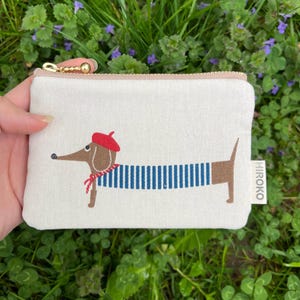 Könnte beinhalten: Eine weiße Canvas-Reißverschlusstasche mit einem niedlichen Cartoon-Dackel, der eine rote Baskenmütze und ein blau-weiß gestreiftes Hemd trägt. Die Tasche hat den Schriftzug "HIROKO" auf der Seite.