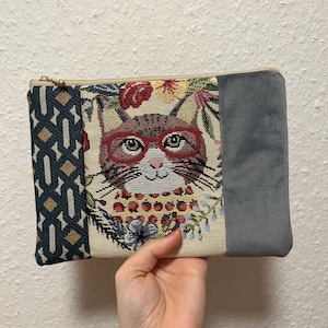 Könnte beinhalten: Rechteckige Clutch mit Katzengesicht-Design. Die Clutch hat einen goldenen Reißverschluss, einen Stoff mit geometrischem Muster, einen Blumenteppich mit einer Brille tragenden Katze und eine graue Samtfläche. Die Katze hat eine rote Brille und ist von Blumen umgeben.