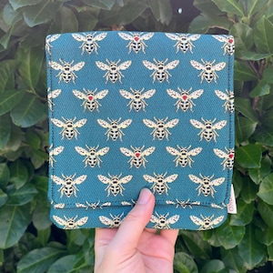 Op de afbeelding: Een vierkant, blauwgroen stoffen tasje met een herhalend patroon van gouden en zwarte bijen. Elke bij is gedetailleerd met een klein, opvallend rood hartje op de rug. Het tasje heeft een klepsluiting en een zichtbaar wit stoffen label aan de zijkant.