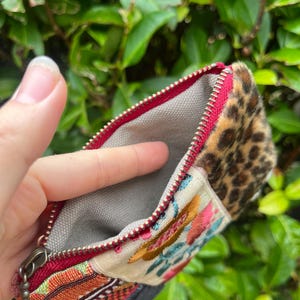 May include: Open zippered pouch with a red zip and a grey interior. The pouch features a section with a leopard print pattern, a section with colourful embroidery, and a section with a woven design.