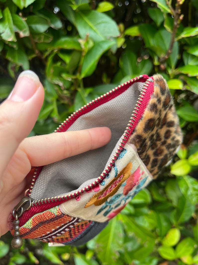 May include: Open zippered pouch with a red zip and a grey interior. The pouch features a section with a leopard print pattern, a section with colourful embroidery, and a section with a woven design.