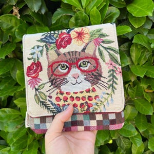 Könnte beinhalten: Ein gewebtes Stoffetui mit einem braunen getigerten Katzenmotiv, das eine rote Brille trägt und von einem Kranz aus roten und gelben Blumen umgeben ist. Der untere Teil des Etuis zeigt ein mehrfarbiges Karomuster in Rot-, Grün- und Blautönen.
