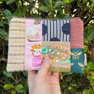 Könnte beinhalten: Eine Patchwork-Reißverschlusstasche mit verschiedenen Stofftexturen und Mustern. Das Design umfasst gestickte Katzen, florale Motive und geometrische Formen in Rosa, Beige, Marineblau und Türkis. Die Tasche hat einen goldenen Reißverschluss und ein kleines Etikett mit "HIROKO".