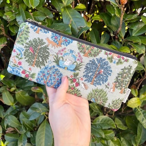 Op de afbeelding: Een rechthoekige etui met een patroon met een natuurthema. De etui heeft een herhalend ontwerp van bomen, vogels, vossen en een zwaan op een lichtgroene achtergrond. De rits is zwart en de etui heeft een klein label met "HIROKO".