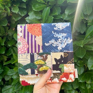 Könnte beinhalten: Eine quadratische Stoffgeldbörse mit einem Patchwork-Design aus verschiedenen Mustern, darunter violette Streifen mit rosa Chrysanthemen, blaue Wellen, grüne Kiefern, eine schwarze Katzensilhouette und rote Blumenmotive mit einem weißen Etikett HIROKO.