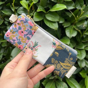 Könnte beinhalten: Eine kleine Reißverschlusstasche mit Patchwork-Design. Die Tasche besteht aus vier verschiedenen Stoffen, die jeweils ein Blumenmuster aufweisen. Die Tasche hat einen goldenen Reißverschluss und einen weißen Riemen mit einem goldenen Ring. Die Tasche ist mit "HIROKO" beschriftet.