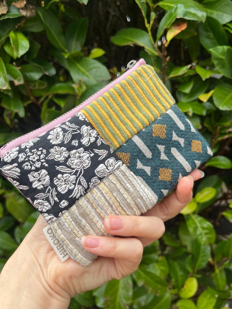 May include: A multi-fabric patchwork pouch with a pink zip. The pouch features a variety of patterns, including black floral, yellow striped, teal geometric, and beige striped designs. The brand name "HIROKO" is visible on a small tag.