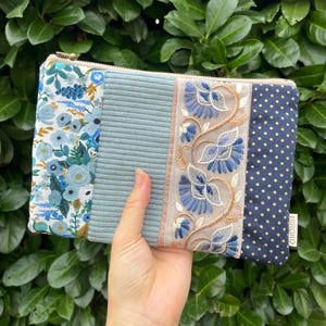 Könnte beinhalten: Eine rechteckige Reißverschlusstasche mit vier verschiedenen vertikalen Paneelen: ein Blumenmuster mit blauen und weißen Blüten, ein hellblauer Kordstoffabschnitt, ein beiges Paneel mit blauer Blumenstickerei und ein dunkelblauer Stoff mit goldenen Punkten.