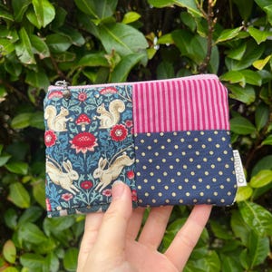 May include: A small, zippered pouch with a patchwork design. One section features a dark blue floral pattern with squirrels and rabbits. Another section has pink vertical stripes, and the third section has navy blue polka dots. The pouch is held in a hand.