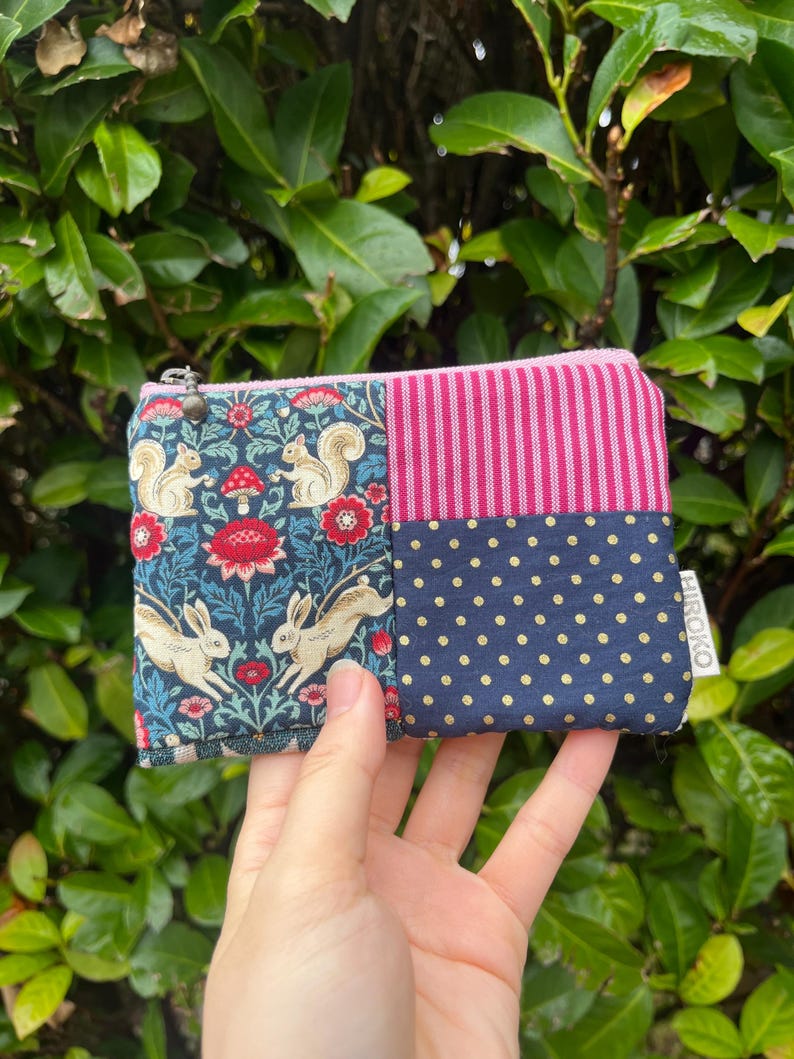 May include: A small, zippered pouch with a patchwork design. One section features a dark blue floral pattern with squirrels and rabbits. Another section has pink vertical stripes, and the third section has navy blue polka dots. The pouch is held in a hand.