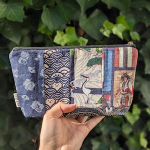 Może przedstawiać: Niebieska i beżowa kosmetyczka w patchworku z różnymi wzorami inspirowanymi kulturą japońską, w tym żurawiami, falami i kotami. Kosmetyczka ma czarny zamek błyskawiczny i małą etykietę z napisem "HIRO".