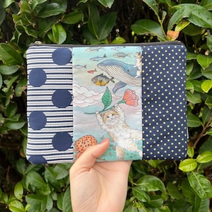 Könnte beinhalten: Eine marineblau und weiß gemusterte Reißverschlusstasche. Die Tasche hat drei Paneele: ein gestreiftes Paneel mit großen marineblauen Kreisen, ein Paneel mit einer skurrilen Katzen- und Walillustration und ein Paneel mit kleinen gelben Punkten.