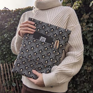 Könnte beinhalten: Eine schwarz-weiß gemusterte Mappe mit einem fächerartigen Design. Die Mappe hat eine Tasche mit einer Visitenkarte und einem Stift. Die Mappe wird von einer Person gehalten, die einen cremefarbenen Pullover trägt.