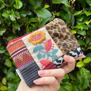May include: A multi-textured clutch bag with a floral embroidered panel featuring a yellow and pink flower, a leopard print section, and a black and white patterned section. The bag has a ribbed grey section.