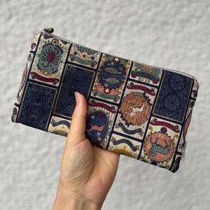 Könnte beinhalten: Ein rechteckiges Reißverschlussetui aus Stoff mit einem wiederkehrenden Muster aus Tarot-Karten-Motiven, die Sternzeichen wie Skorpion, Fische und Steinbock zeigen. Das Design verwendet eine Palette aus Marineblau, Creme und Erdtönen.