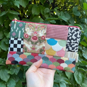 Könnte beinhalten: Eine Patchwork-Clutch mit rotem Reißverschluss und goldenem Zug. Das Design beinhaltet verschiedene Muster, darunter ein schwarz-weißes Schachbrettmuster, Blumendrucke mit Eichhörnchen und Kaninchen sowie einen strukturierten rosa Streifen. Der Boden hat ein farbenfrohes, schuppenartiges Muster.