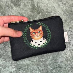 Könnte beinhalten: Schwarzes Reißverschlussetui mit einer skurrilen Katzenillustration. Die Katze mit orangefarbenem Fell und Brille ist von einem grünen Kranz umgeben und trägt einen gepunkteten Kragen. Das Etui hat einen goldenen Reißverschluss und ein kleines "HIROKO"-Etikett.