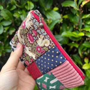 May include: A patchwork zipped pouch with a variety of fabric patterns, including squirrels, floral designs, polka dots, and corduroy. The pouch has a red zip and trim, and a small metal zip pull.