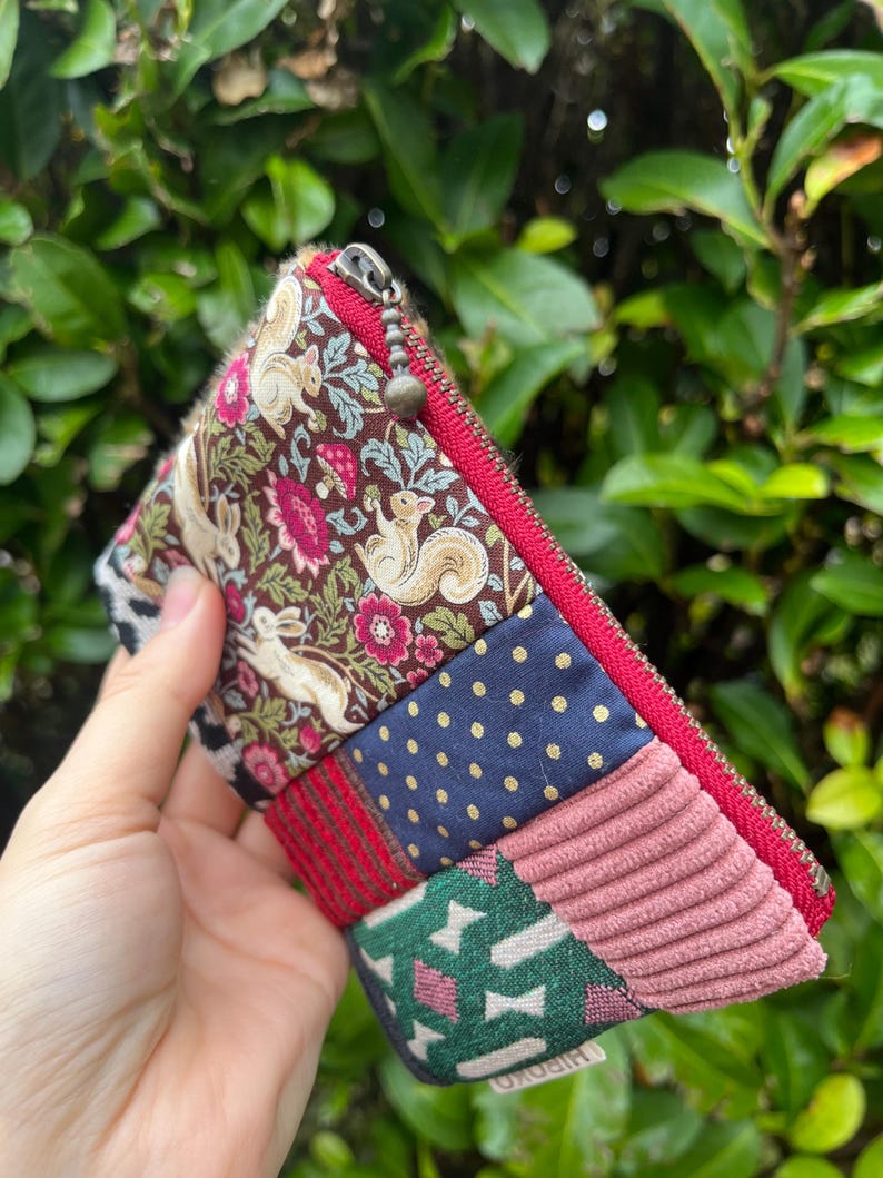 May include: A patchwork zipped pouch with a variety of fabric patterns, including squirrels, floral designs, polka dots, and corduroy. The pouch has a red zip and trim, and a small metal zip pull.