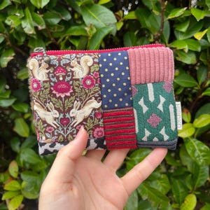 Könnte beinhalten: Kleine, patchworkartige Reißverschlusstasche mit verschiedenen gemusterten Stoffen. Die Tasche hat einen floralen und tierischen Druck, marineblaue Polka Dots, rosa Cord und ein grünes geometrisches Design. Der Reißverschluss ist rot.