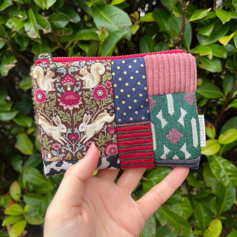 May include: A small, patchwork zipped pouch with various patterned fabrics. The pouch features a floral and animal print, navy blue polka dots, pink corduroy, and a green geometric design. The zip is red, and the pouch is held in a hand.