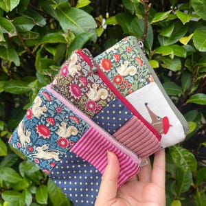May include: A selection of small fabric pouches with zip closures. The pouches display diverse patterns, such as floral, polka dots, and animal motifs. The colour scheme includes blue, pink, red, and green. The pouches are held in a hand.