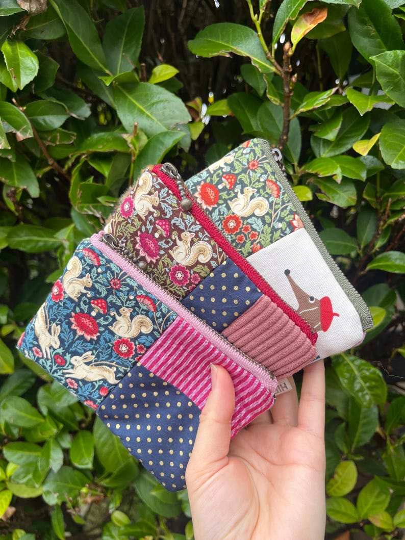May include: A selection of small fabric pouches with zip closures. The pouches display diverse patterns, such as floral, polka dots, and animal motifs. The colour scheme includes blue, pink, red, and green. The pouches are held in a hand.
