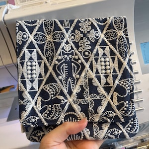Op de afbeelding: Een donker marineblauw stoffen tasje met een ingewikkeld wit geborduurd ruitpatroon. Het ontwerp bevat gestileerde motieven van katten, vogels en geometrische vormen in elke ruit. Het textiel heeft een zichtbare weving en een klepsluiting.