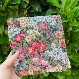 Könnte beinhalten: Eine quadratische Stofftasche mit einem gewebten Gobelin-Muster aus bunten Blumen, darunter rote, gelbe und hellblaue Blüten auf dunkelgrünem und schwarzem Grund. Die Tasche hat einen Klappverschluss und ein kleines weißes Etikett an der Seite.