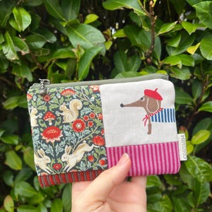 May include: A small, zipped coin purse with a patchwork design. One side has a floral pattern with squirrels and rabbits, the other a dog in a beret and striped shirt. The purse is held in a hand.