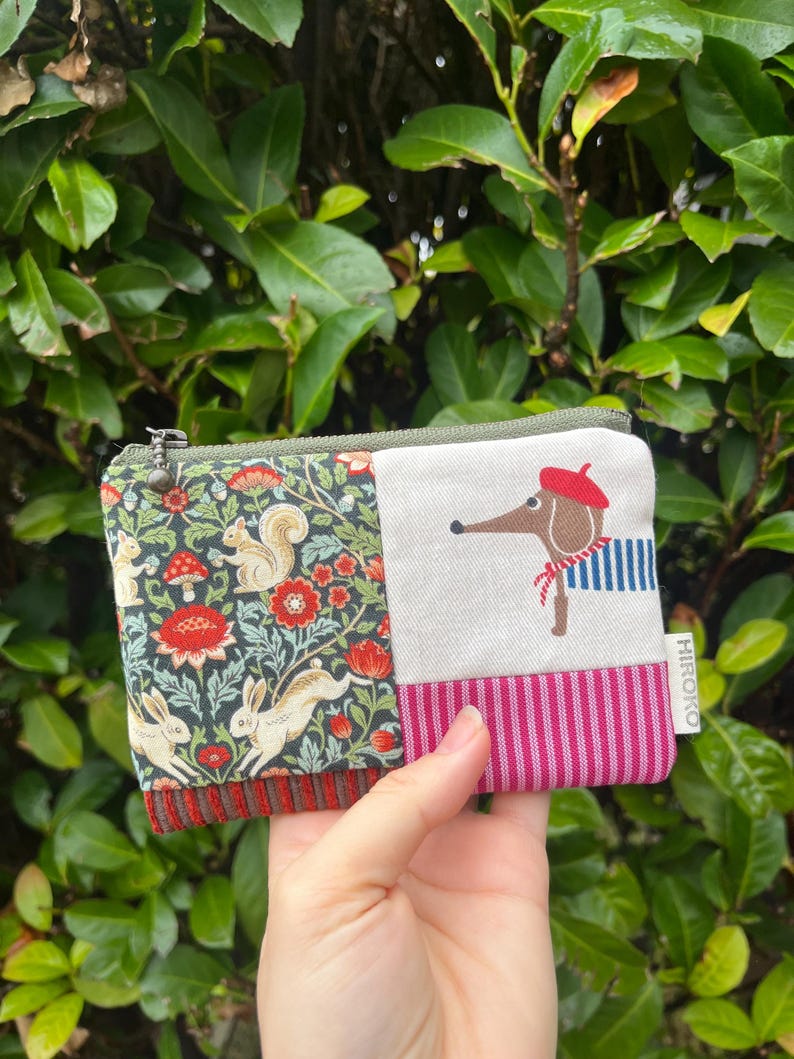 May include: A small, zipped coin purse with a patchwork design. One side has a floral pattern with squirrels and rabbits, the other a dog in a beret and striped shirt. The purse is held in a hand.