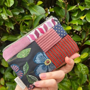 May include: A multi-fabric patchwork wallet with a variety of patterns and textures. The wallet features a floral design, stripes, polka dots, and solid colours. The label reads "HIROKO".