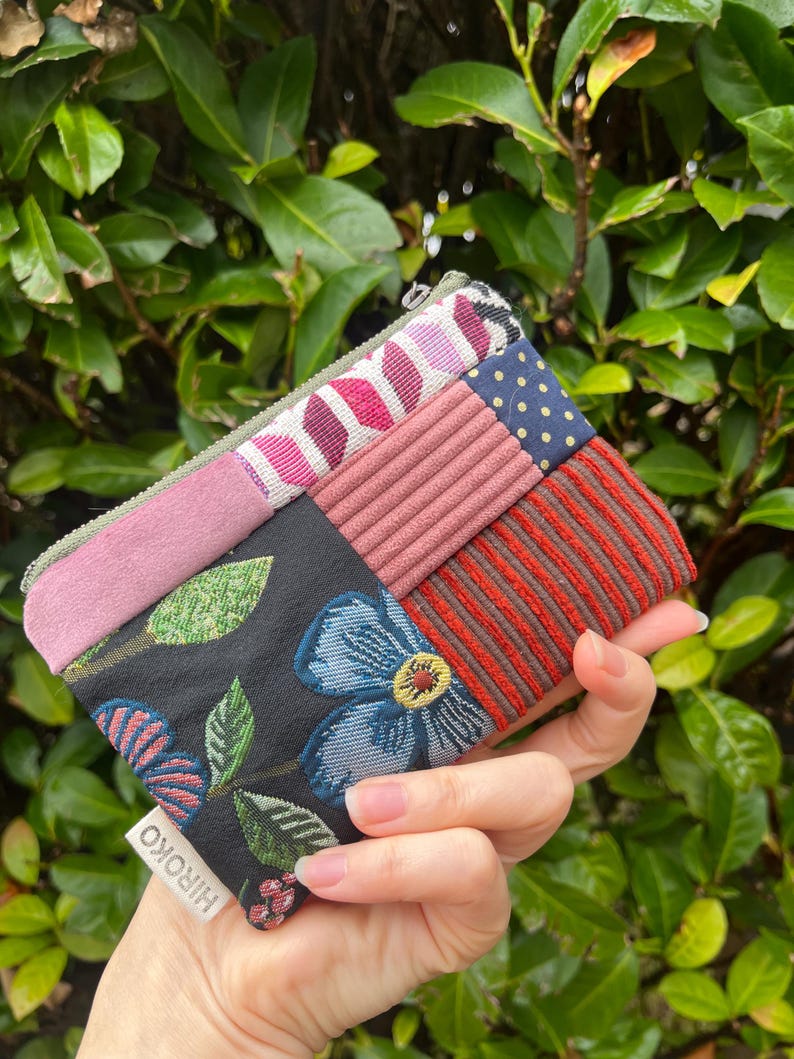 May include: A multi-fabric patchwork wallet with a variety of patterns and textures. The wallet features a floral design, stripes, polka dots, and solid colours. The label reads "HIROKO".