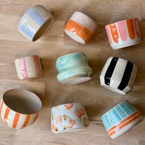 Könnte beinhalten: Eine Sammlung handgefertigter Keramikbecher mit verschiedenen Designs. Die Becher weisen Farben wie Orange, Rosa, Blau und Grün mit gestreiften und abstrakten Mustern auf. Die Becher sind auf einer Holzoberfläche angeordnet.