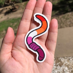 May include: A sticker of a worm with a lesbian pride flag color scheme. The worm is pink, orange, white, and red. The sticker has a black outline and the text "@juicy.wear" is visible on the sticker.