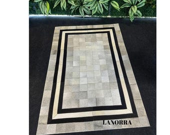 May include: A white and gray cowhide rug with black and white trim. The rug has a square pattern and the text "LANORRA" is printed in the bottom right corner.