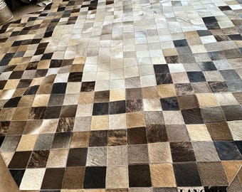 May include: A large, geometric area rug composed of squares in shades of brown, beige, and black. The rug's design features a patchwork pattern, creating a visually interesting texture. The rug is made of a natural material, likely hide or leather.