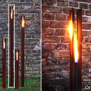 Könnte beinhalten: Vier hohe, rostige Metall-Fackelhalter mit brennenden Flammen im Inneren. Die Fackeln stehen vor einer Backsteinmauer.