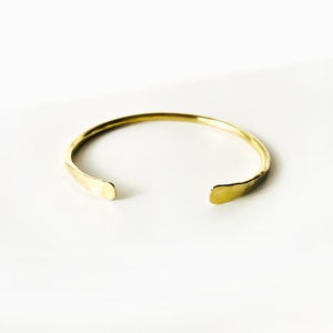 Könnte beinhalten: Ein goldfarbenes, gehämmertes Metall-Armreif-Armband. Das Armband ist offen und hat ein schlichtes, minimalistisches Design.