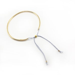 Może przedstawiać: Złota bransoletka z delikatnym, regulowanym zapięciem na sznurek. Sznurek jest jasnoniebieski i ma małą złotą kulkę w środku.