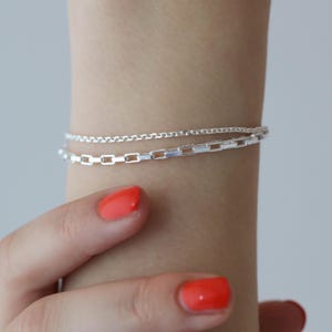 May include: Two silver bracelets are shown on a wrist. The top bracelet is a delicate chain, and the bottom bracelet features rectangular links. The person's nails are painted with bright orange nail polish.