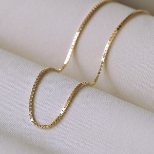 Op de afbeelding: Een delicate goudkleurige ketting met een box chain design. De ketting is gemaakt van kleine, vierkante schakels die het licht vangen. De ketting wordt gepresenteerd op een witte, getextureerde stof, wat een eenvoudige en elegante presentatie creëert.