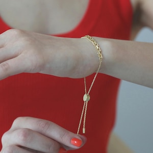 Puede incluir: Pulsera dorada con un delicado diseño de cadena. La pulsera presenta un cierre deslizante ajustable y se muestra en una muñeca sobre un fondo rojo. El diseño de la pulsera incluye pequeños eslabones rectangulares.