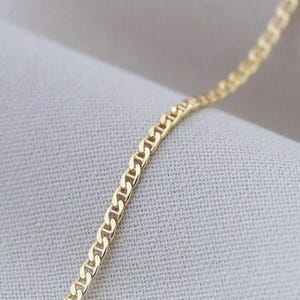 Könnte beinhalten: Nahaufnahme einer goldenen Halskette mit Ankerketten-Design. Die Kette wird auf einem neutralfarbenen Stoff präsentiert, der ihre glänzende, gelbgoldene Farbe hervorhebt. Die Glieder sind gleichmäßig angeordnet und erzeugen ein strukturiertes Muster.