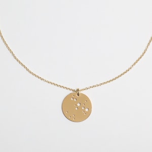May include: A gold necklace with a fine chain and a circular pendant. The pendant features a constellation design with small, evenly spaced cut-out holes. The necklace is set against a plain white background.