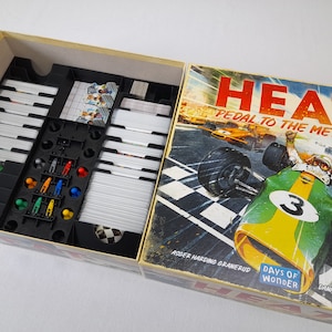 Puede incluir: Una caja de juego de mesa con un coche de carreras verde y amarillo en la portada. La caja dice "HEAT Pedal to the Metal" y tiene el logotipo de Days of Wonder. La caja está abierta y muestra los componentes del juego en el interior, incluyendo cartas, fichas y un tablero de juego.