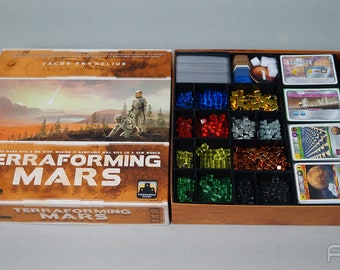 May include: A Terraforming Mars board game box with a brown and orange design. The box features a picture of a red planet with a lake and two astronauts. The text "Terraforming Mars" is printed in white on the box. The box is open and shows the game components, including cards, dice, and tokens.