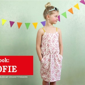 Op de afbeelding: Een meisje draagt een roze-witte stippenjurk met spaghettibandjes en zakken. De jurk heeft een getailleerde taille. Het meisje draagt een zonnebril en heeft haar haar in een knotje geknoopt. De achtergrond is een lichtgroene muur met een kleurrijke papieren slinger.