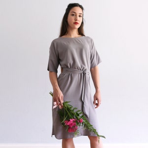 May include: A gray midi dress with a self-tie belt and short sleeves. The dress is made of a soft, flowing fabric and has a relaxed fit. The model is holding a bouquet of pink flowers.