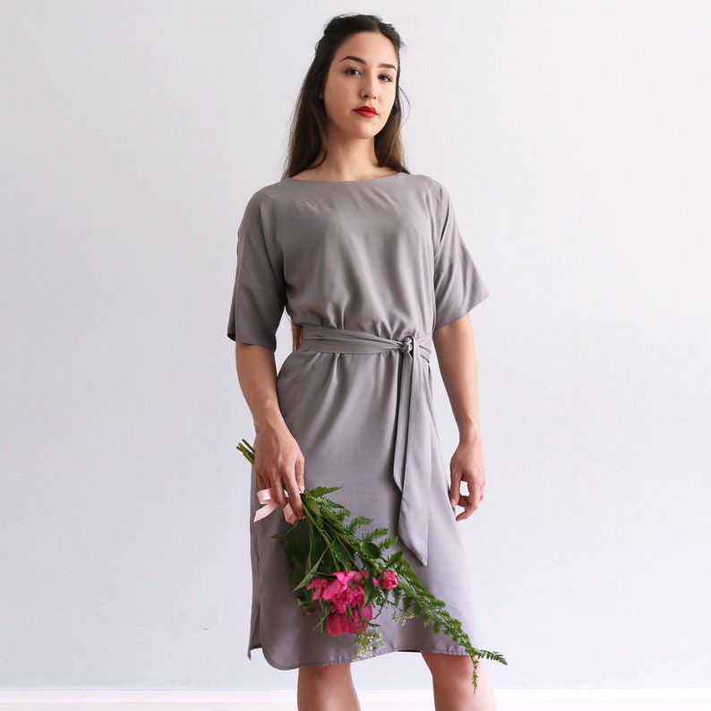 May include: A gray midi dress with a self-tie belt and short sleeves. The dress is made of a soft, flowing fabric and has a relaxed fit. The model is holding a bouquet of pink flowers.